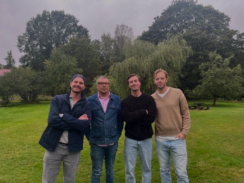 A man and a woman are cleaning a bright, modern room. The woman is holding a mop while wearing pink rubber gloves and looking at a shelf. The man, also in casual attire, is arranging items on the shelf while wearing yellow rubber gloves. There are plants and decorative items on the shelves, and a vibrant abstract painting is displayed on the wall.