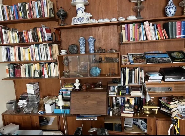 A young woman wearing a white t-shirt and yellow cleaning gloves is wiping a window with a cloth, smiling as she enjoys the sunlight streaming in. Green plants are visible in the foreground.